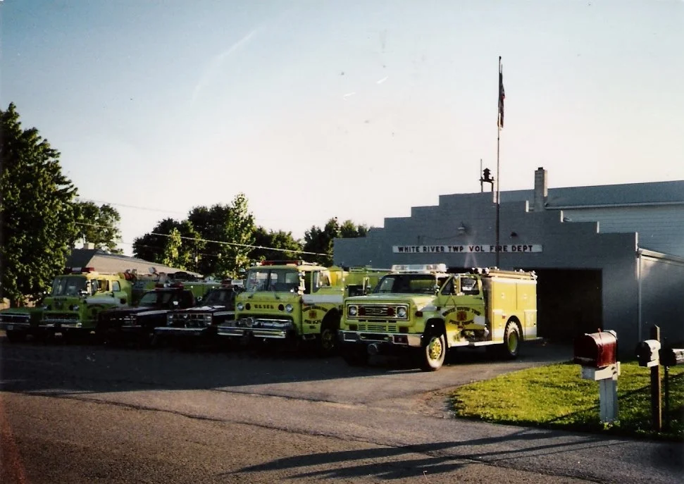Several fire trucks parked outside the White River Twp Volunteer Fire Department building during daytime, with an American flag flying on a pole.