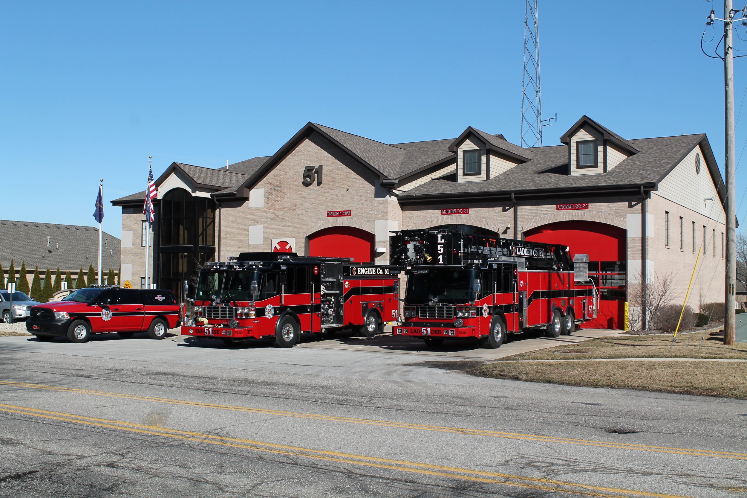 Two red fire trucks parked in front of a fire station building with the number 51 on it. The station has two large red garage doors. A fire chief's vehicle is parked to the left of the fire trucks. The sky is clear and blue.