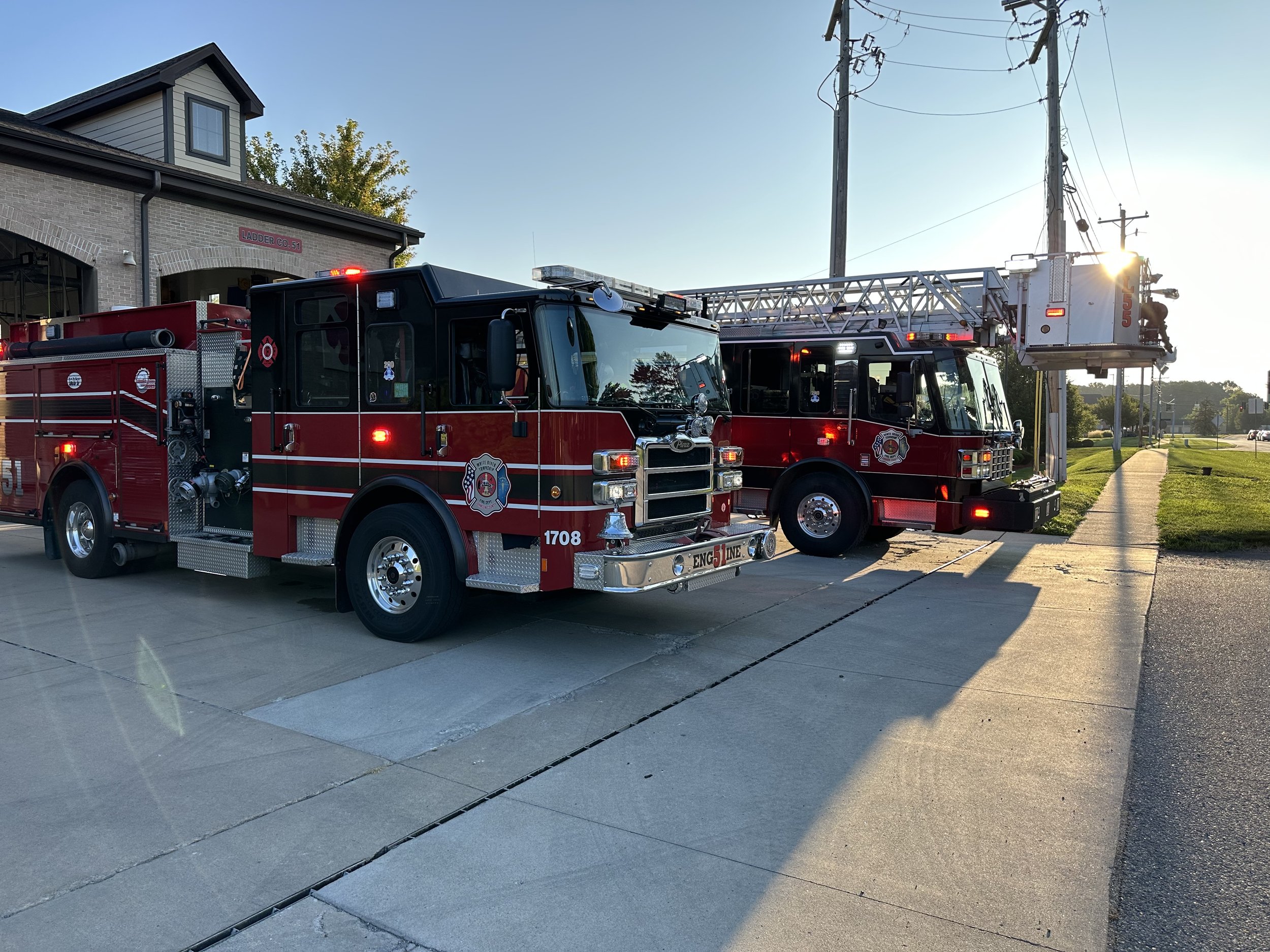 Two red fire trucks parked on a driveway near electric poles, with a building and green trees in the background at sunset.