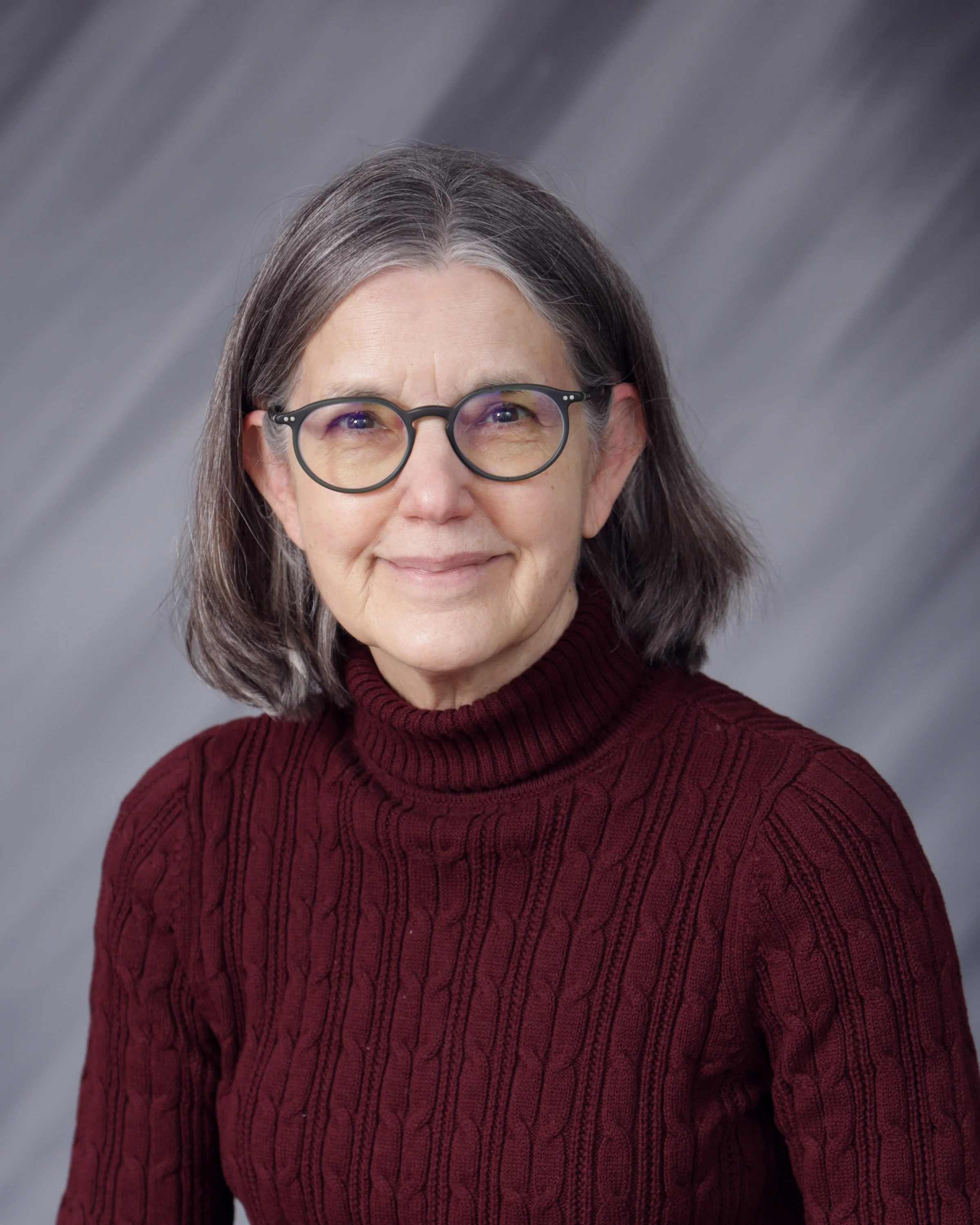 Portrait of an older woman with gray hair, wearing glasses and a maroon turtleneck sweater, smiling softly with a neutral gray background.