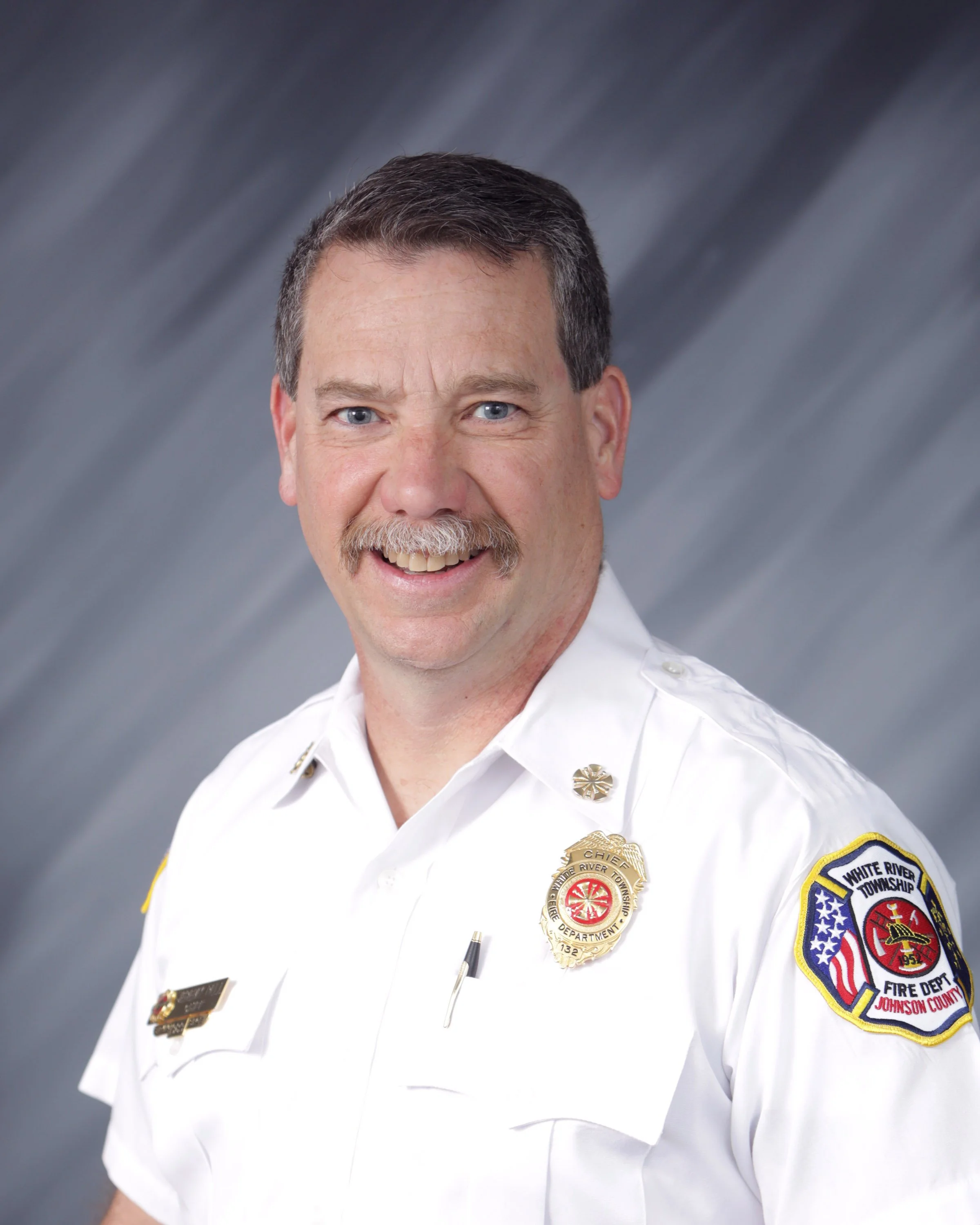 Portrait of a male firefighter in uniform with badges, smiling, against a gray background.