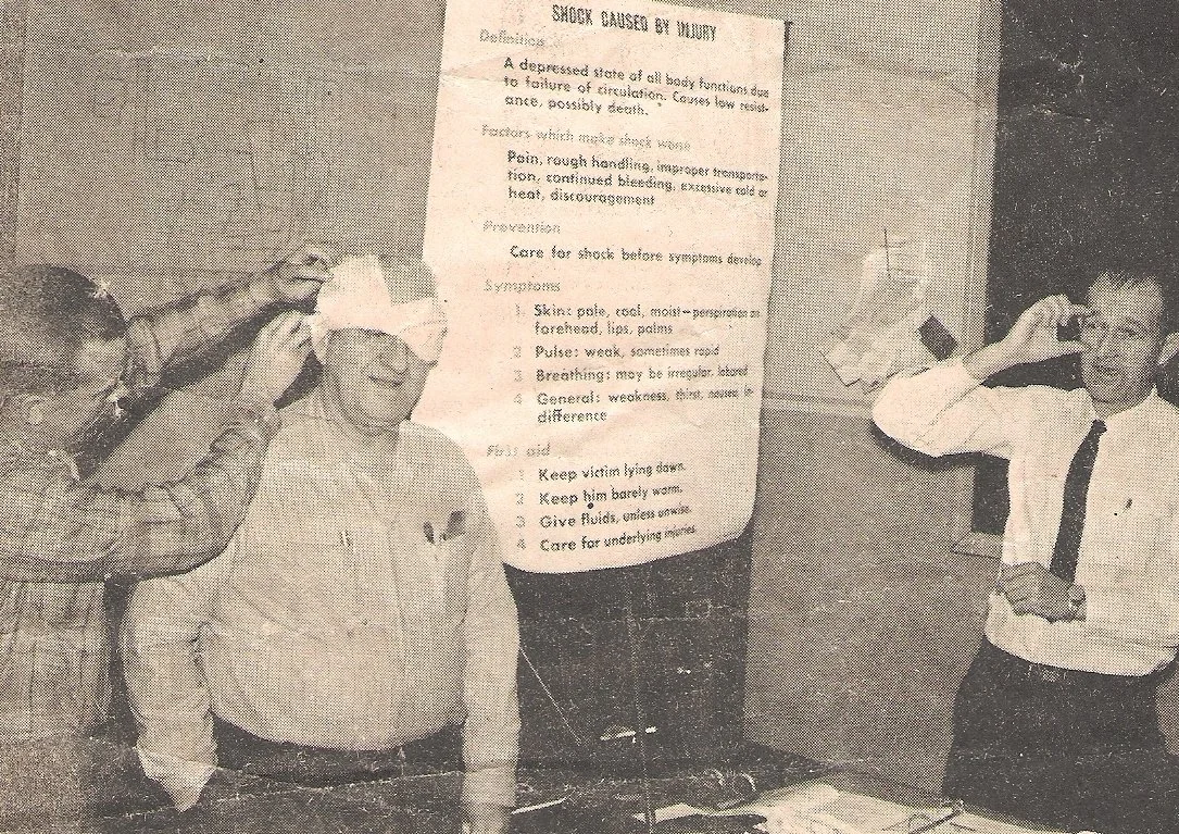 Two men are giving first aid to a third man who has an oxygen mask on, while a fourth man talks on a mobile phone. A poster about shock caused by injury is in the background.