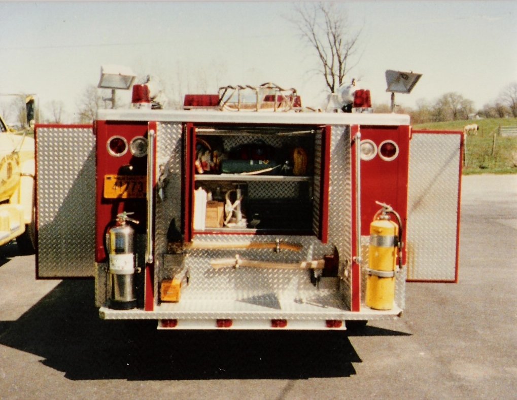 The back of a red fire truck with open compartments showing firefighting equipment, including two fire extinguishers, lights on top, and various tools and supplies inside.