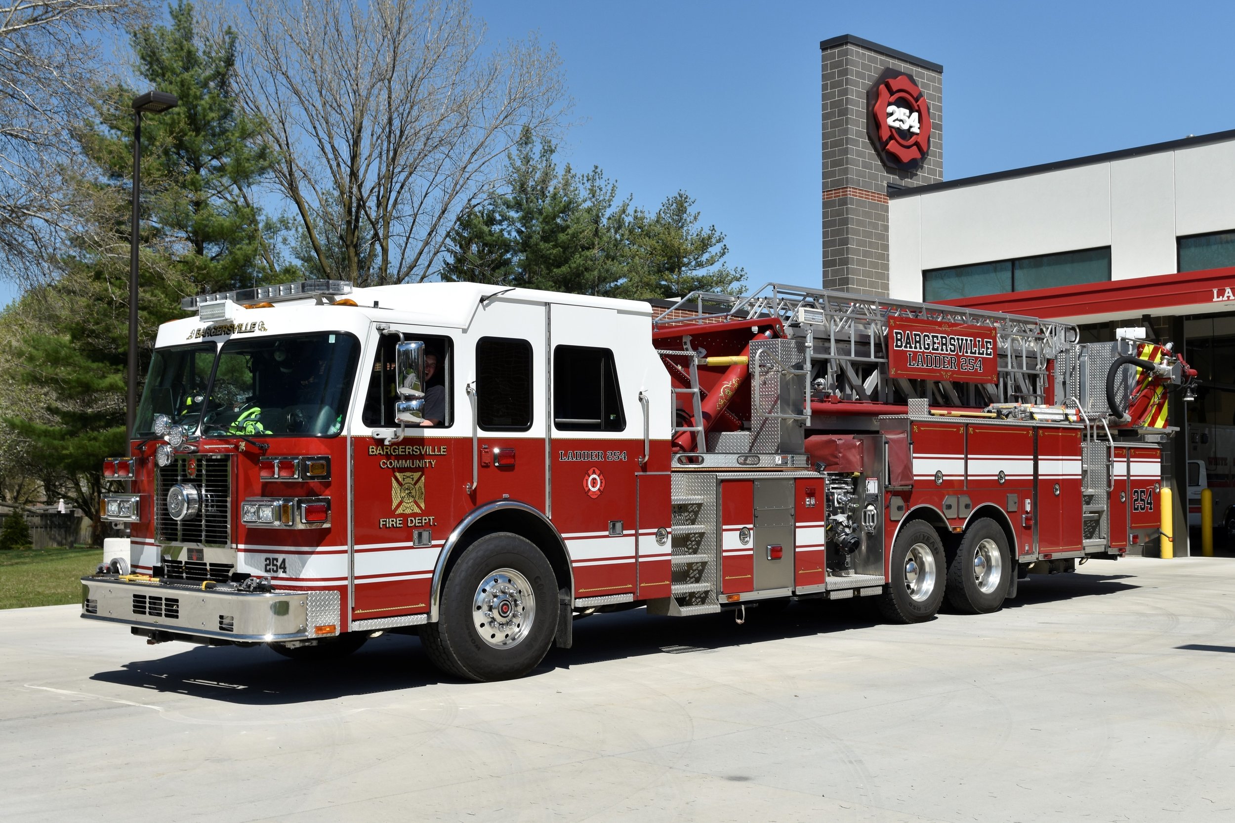 Red and white fire truck parked outside a fire station with a blue sky and trees in the background.