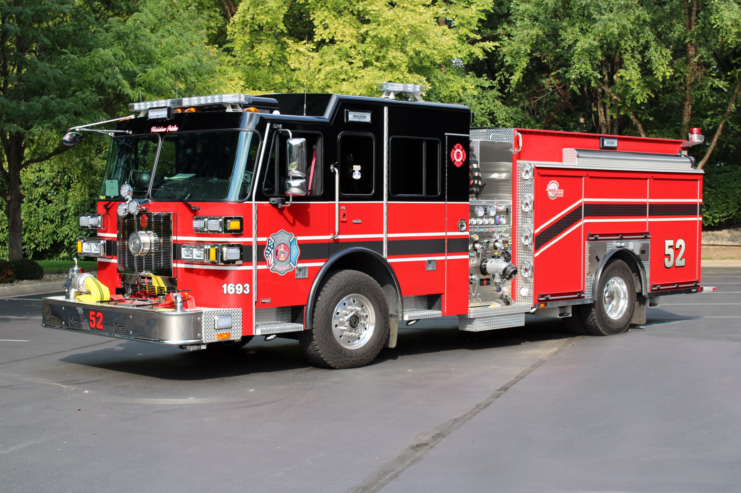 Red fire truck with black and white accents parked on pavement, surrounded by green trees.