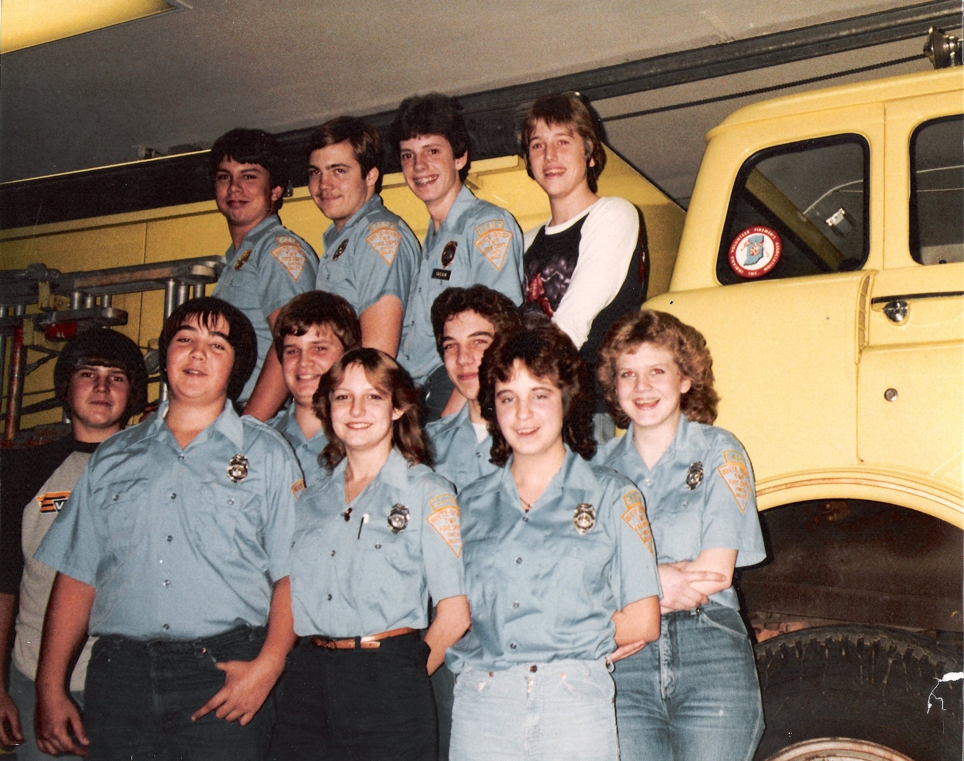 Group of young firefighters and a few girls posing in front of a yellow fire truck, wearing light blue uniforms and looking at the camera.