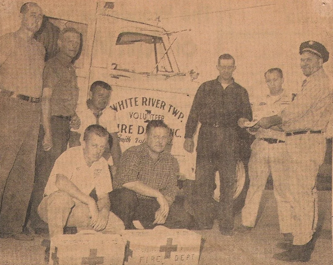 A group of seven people posing in front of a truck with a sign that reads "White River Twp. Youth". Two people are seated in front, and five are standing, including a person in a baseball uniform. One person is holding a book or notebook.