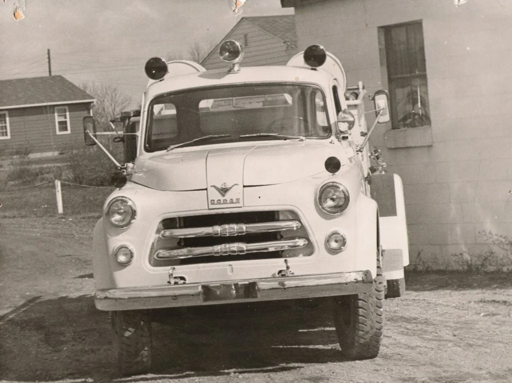 Black and white photo of a vintage vehicle, possibly a fire truck, with a badge that reads 'Dane' on the front. The vehicle has large, prominent headlights and side mirrors. It is parked on a dirt surface near a building with a window, with residenti