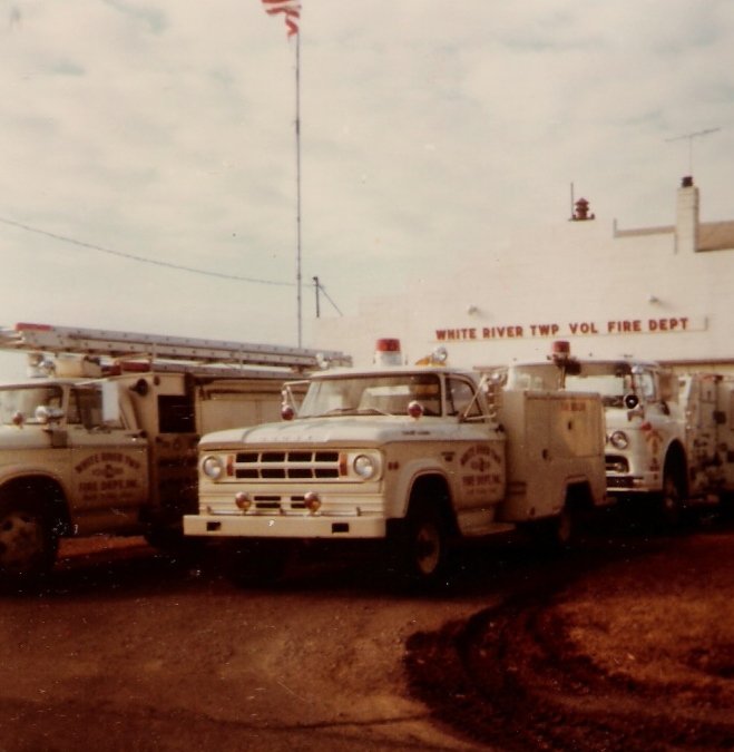 Vintage fire trucks parked outside the White River Township Volunteer Fire Department building, with an American flag flying overhead.