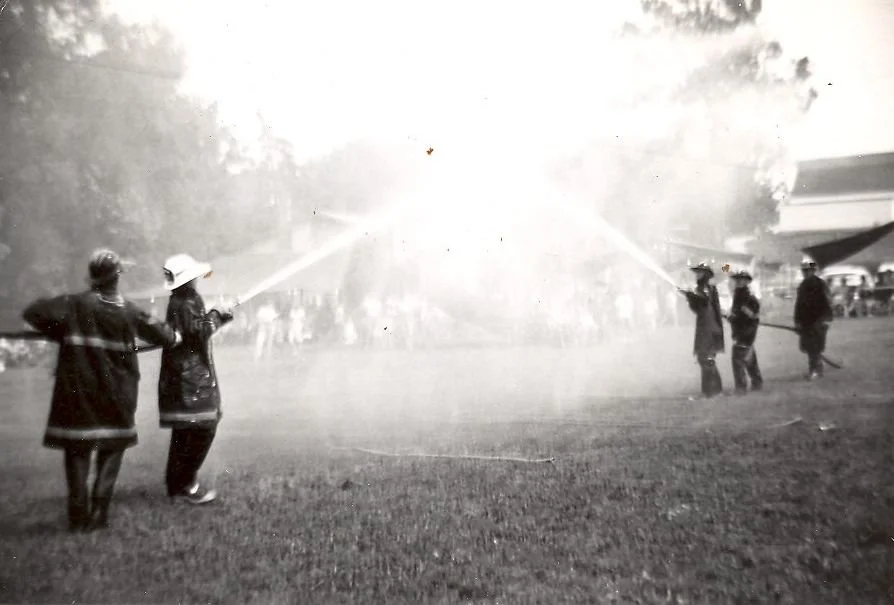 Firefighters spraying water on a fire in an outdoor area, with a crowd in the background.