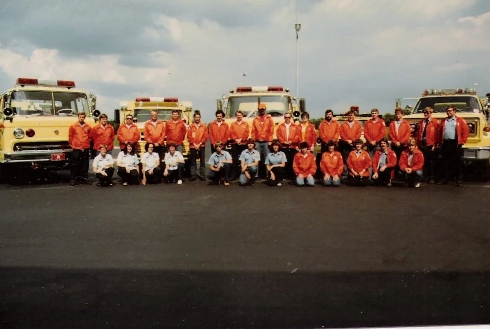 Group of firefighters standing in front of fire trucks, posing for a team photo at a fire station.