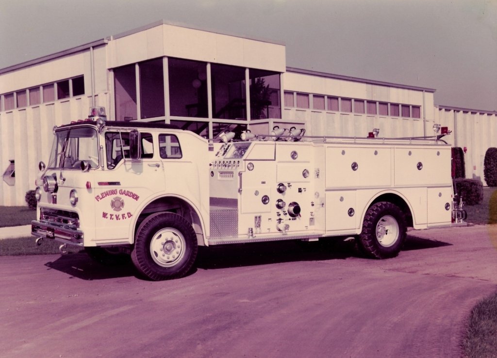 A vintage fire truck parked outside a mid-century modern building, with door lettering reading 'Fleming Garden, K.V.F.D.'