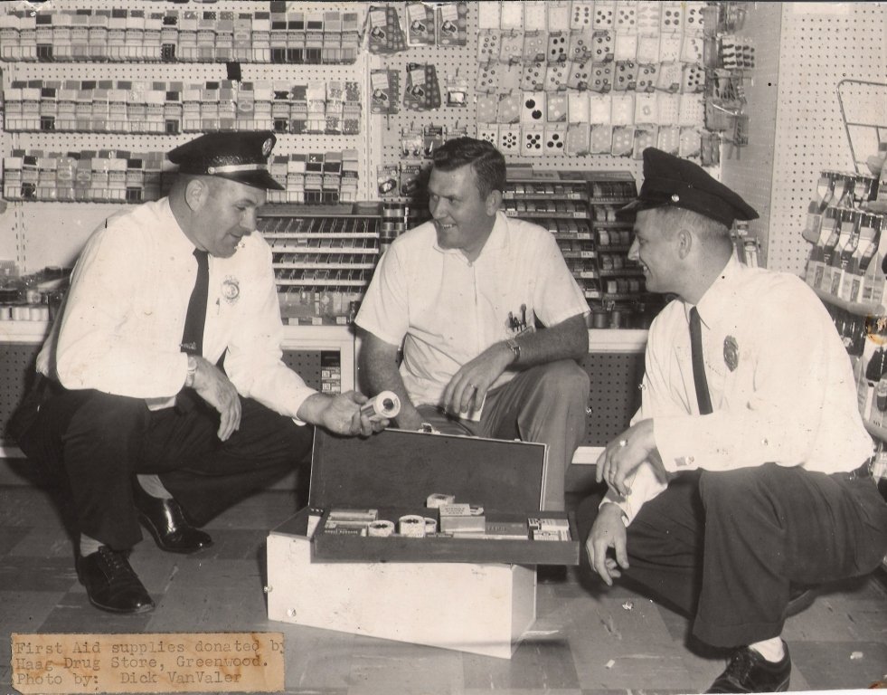 Three men, two police officers and a civilian, sitting on the floor of a drug store surrounded by shelves of supplies. The police officers are in uniform, wearing hats and ties, and appear to be examining first aid supplies donated by the drug store,