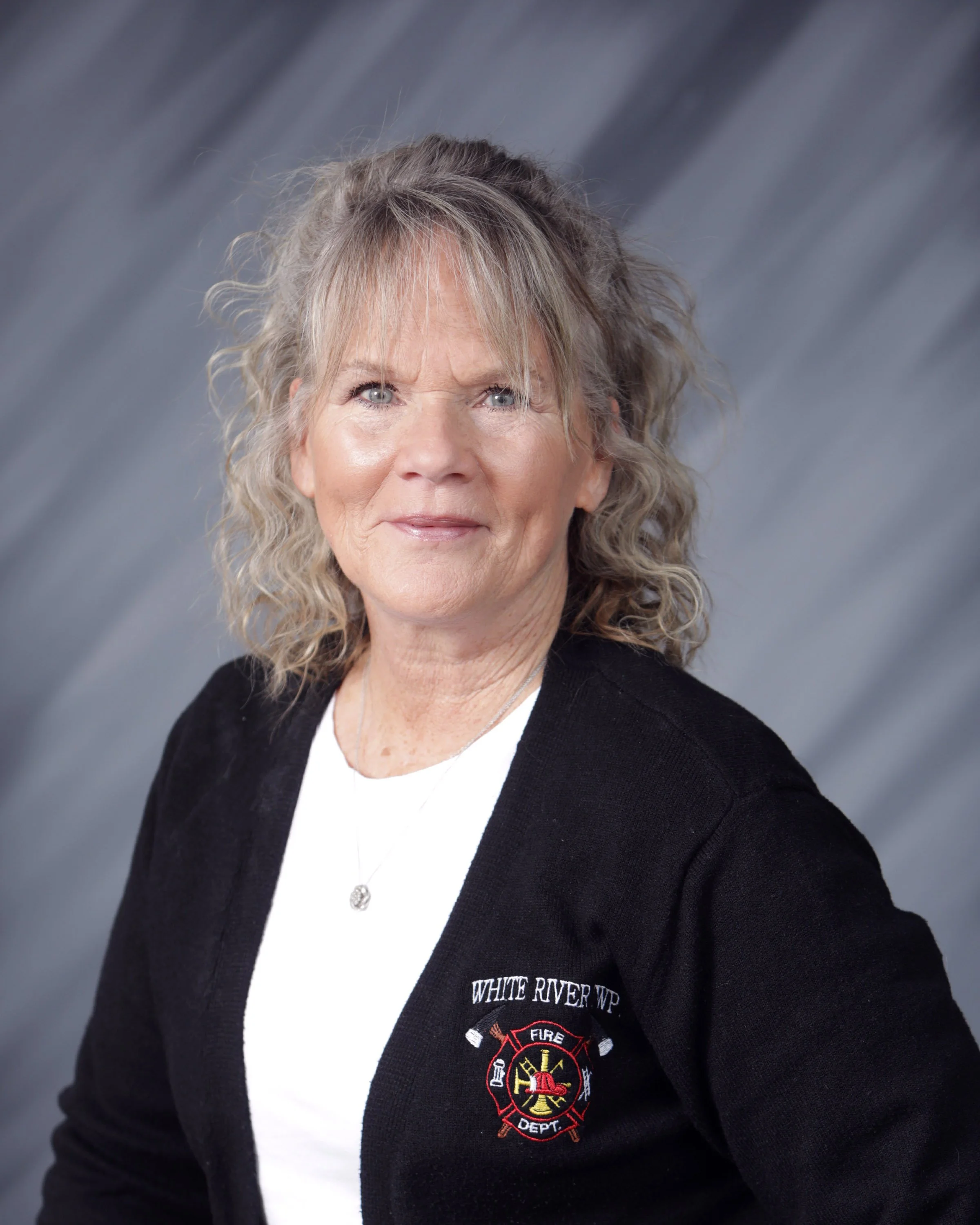 Portrait of a middle-aged woman with curly blonde hair, wearing a black cardigan with an embroidered Fire Department emblem, off-white shirt, and a necklace against a gray background.