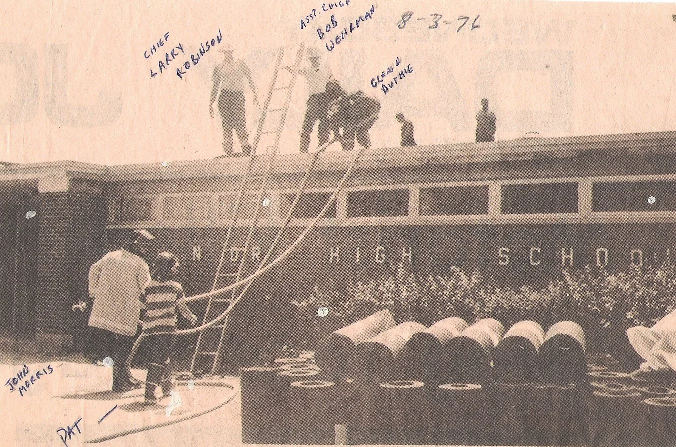 Black and white photo of people working on the roof of a high school building labeled 'High School.' Two individuals are on the ground near a ladder, assisting with work on the roof. Several workers are on the roof, with handwritten notes identifying