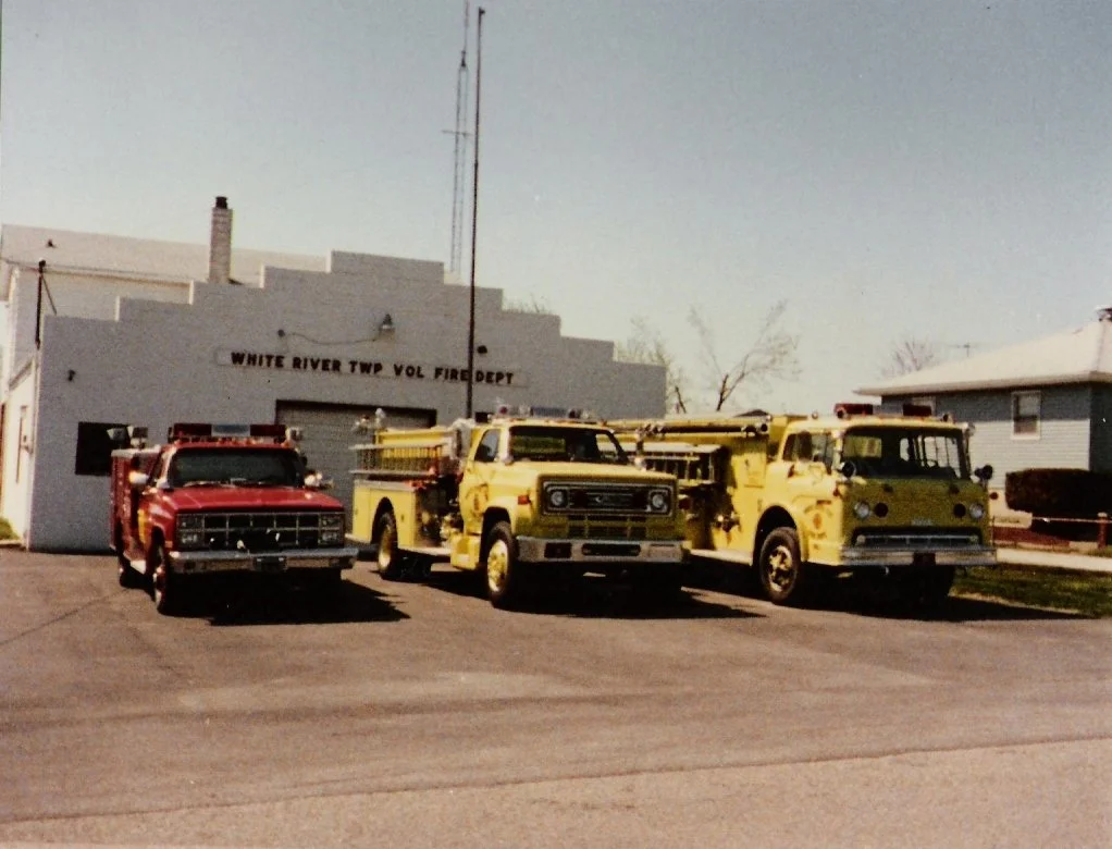 Three vintage fire trucks parked in front of a fire station building labeled 'White River Twp Vol Fire Dept' on a clear day.