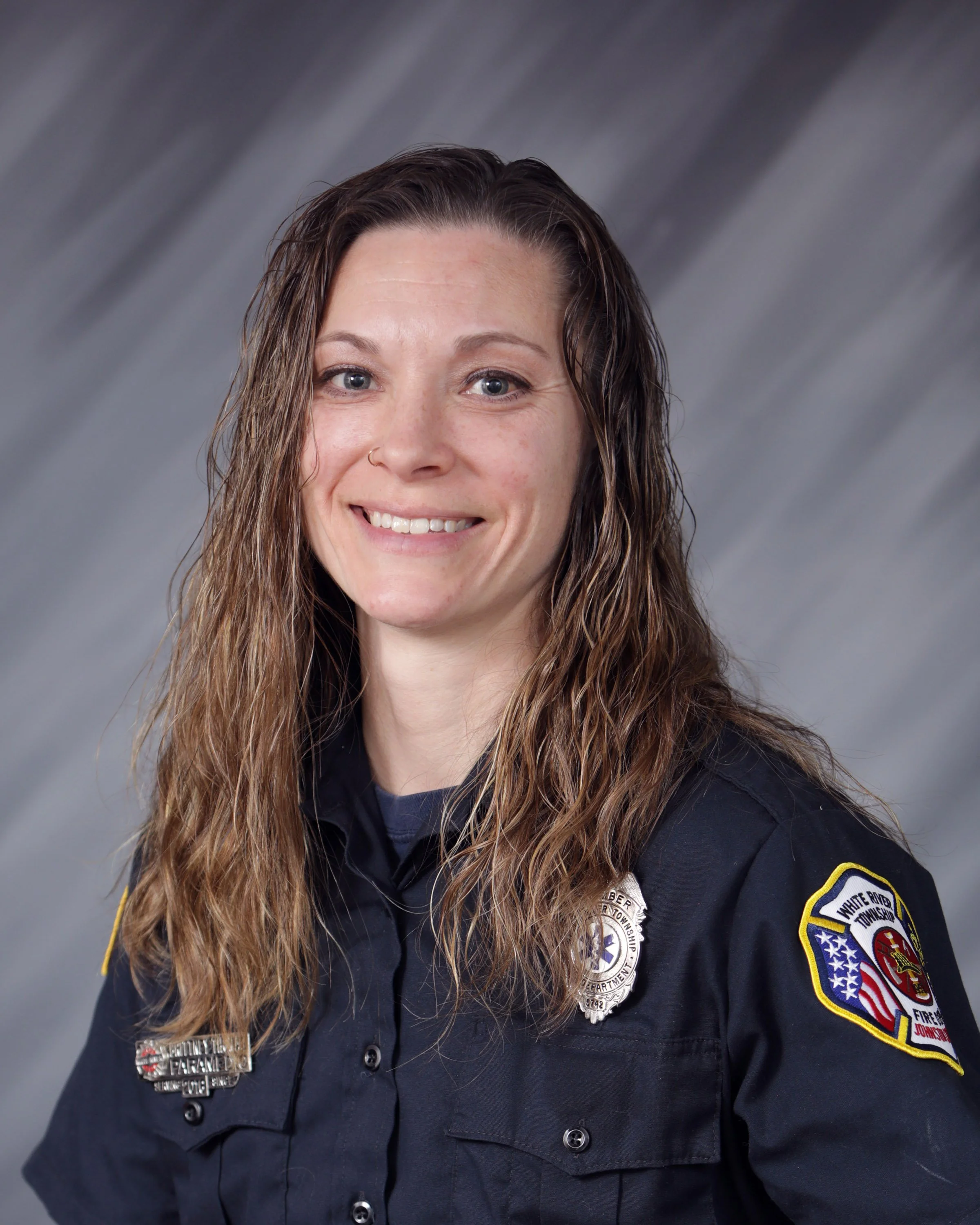 Portrait of a female firefighter with long, wavy brown hair wearing a navy blue uniform with patches and badges, smiling against a gray background.