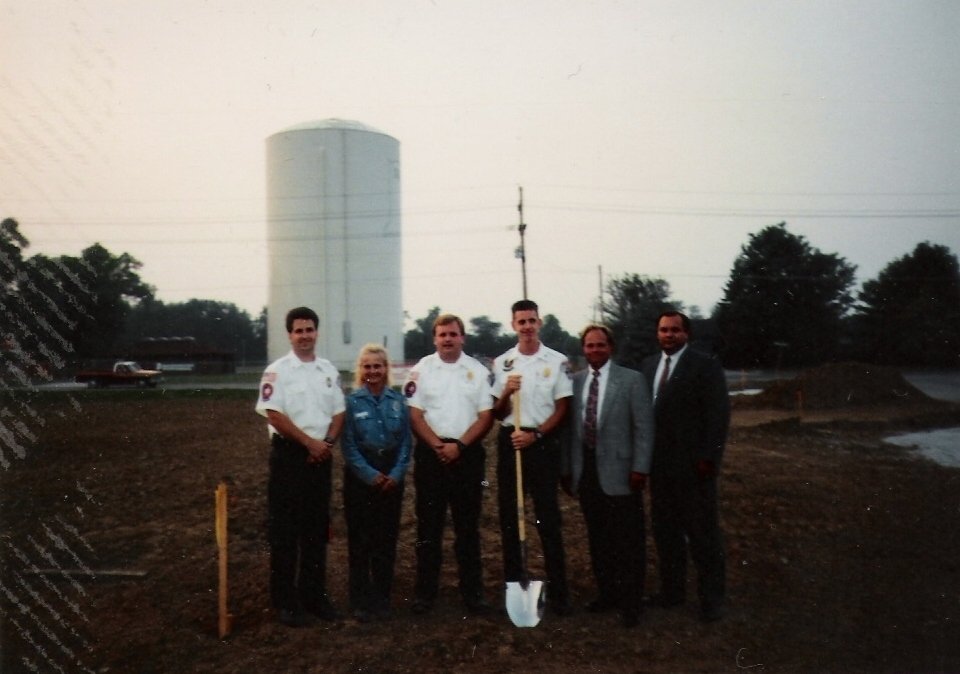 Group of six people, including firefighters and officials, standing on dirt ground during groundbreaking ceremony in front of a large water tower.