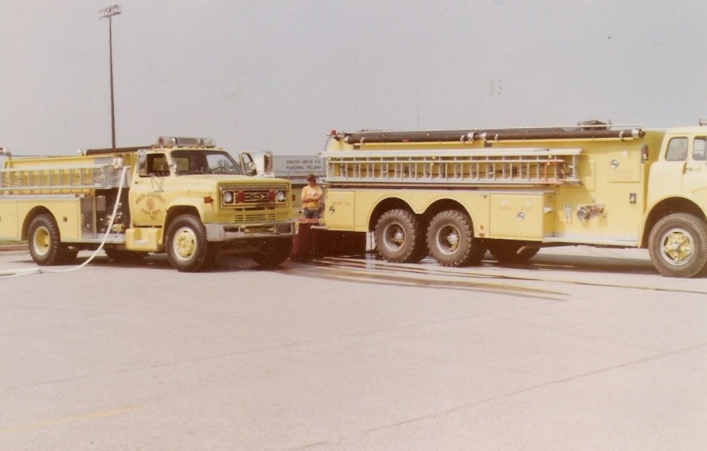Two yellow fire trucks parked on a paved surface with a firefighter standing between them, connecting hoses.