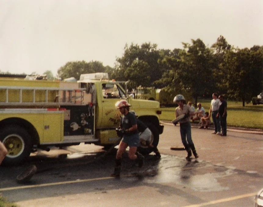 Emergency responders working near a yellow fire utility truck in a parking lot during the daytime with trees and a park nearby, and people sitting and standing in the background.