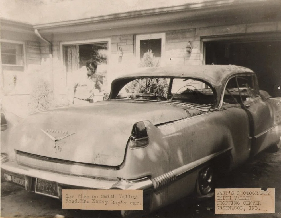 Black and white photograph of a vintage car damaged by fire, parked in front of a house. An older man is standing next to it, looking at the vehicle. The photo is from Smith Valley in Greenwood, Indiana, with notes about a car fire on Smith Valley Ro