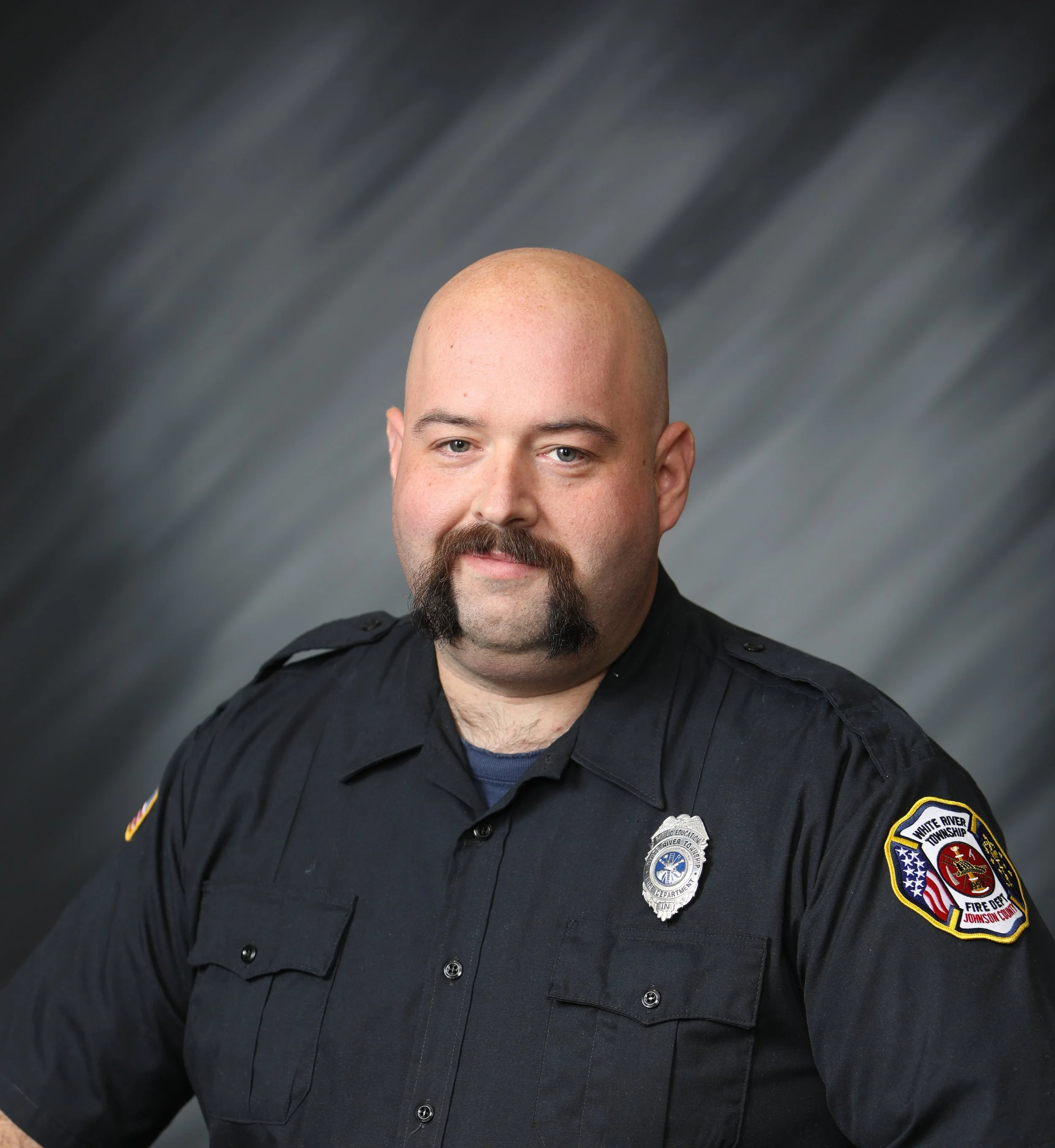 Portrait of a firefighter with a shaved head, mustache, and beard, wearing a uniform with badges, standing against a blurred gray background.
