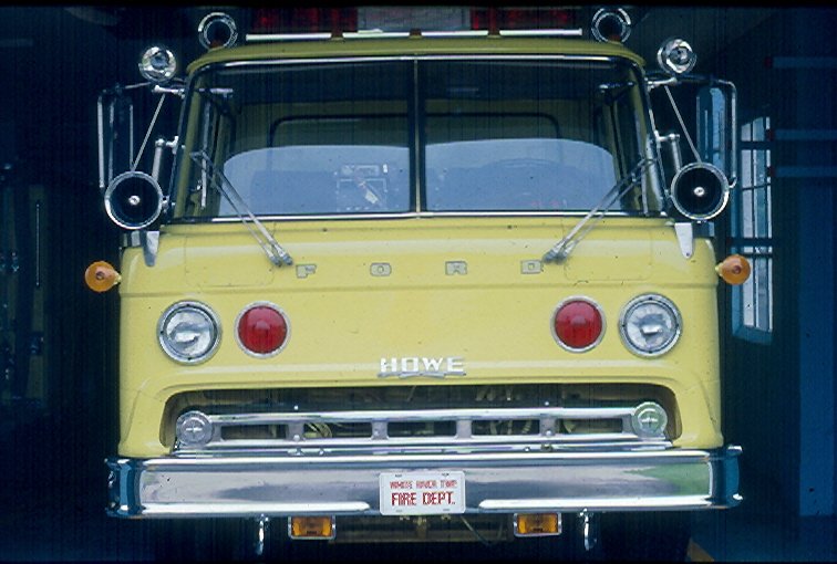 Yellow vintage Ford fire truck with sirens and equipment, parked inside a fire station.