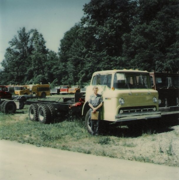 Vintage photograph of a person standing next to a yellow flatbed truck with a blue shirt, parked on grass with trees and other trucks in the background.