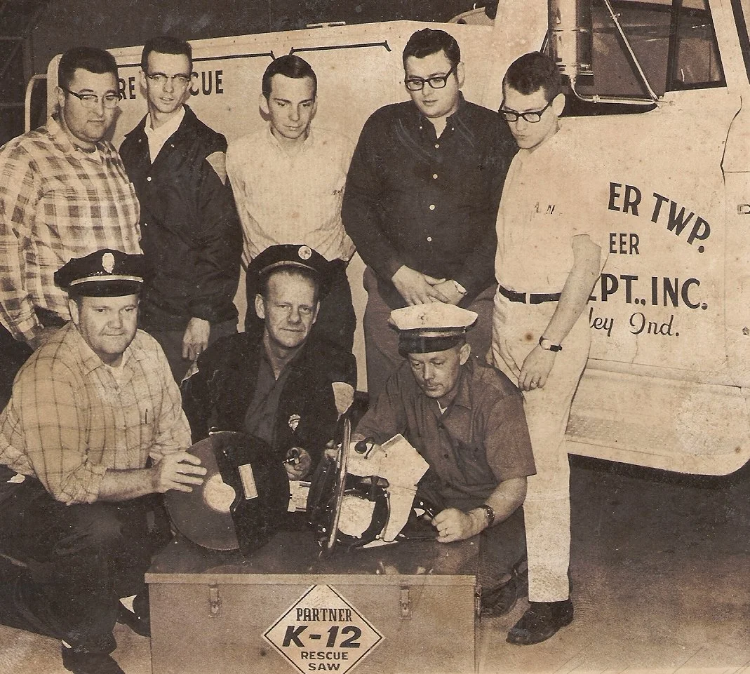 Black and white photo of nine men, some in uniform and some in civilian clothing, posing around rescue equipment including a K-12 rescue saw. A truck with partially visible text is behind them.