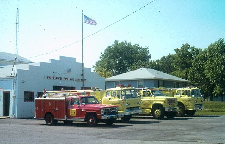 A row of vintage yellow fire trucks and a red fire truck parked outside a fire station, with a building labeled 'White River Twp Vol Fire Dept' and the American flag flying above.