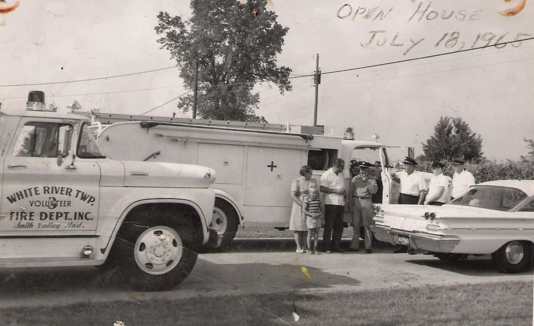 A black and white photo of a group of people, including firemen in uniform and a woman with children, standing near a vintage fire truck, a classic car, and a pickup truck from the 1960s in an outdoor setting.