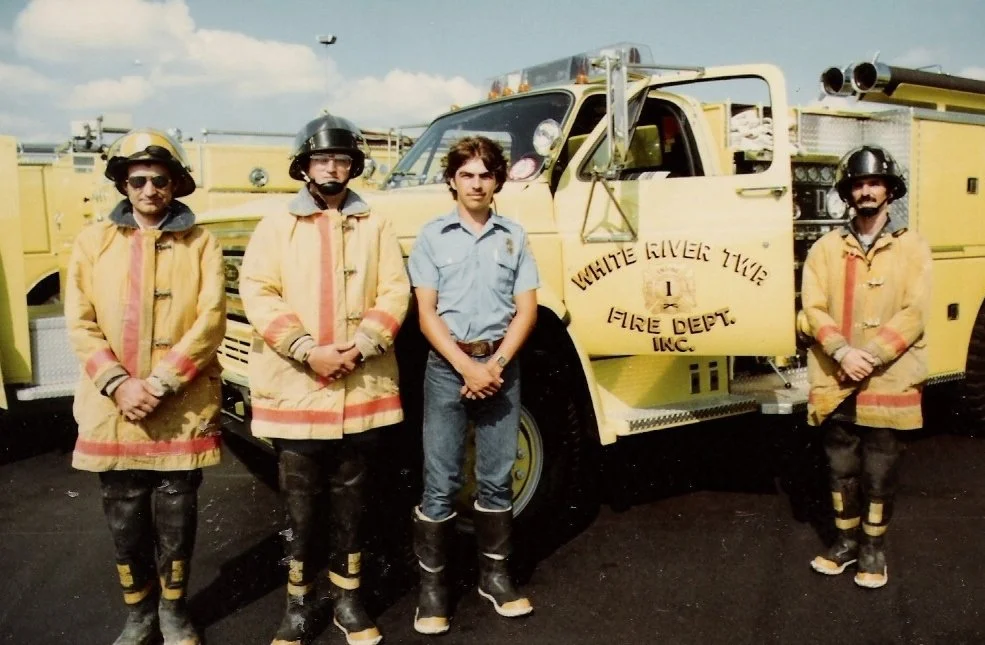 Four firefighters standing in front of a yellow fire truck. One person is dressed in casual clothes, while the others wear firefighting gear, including helmets and protective jackets.