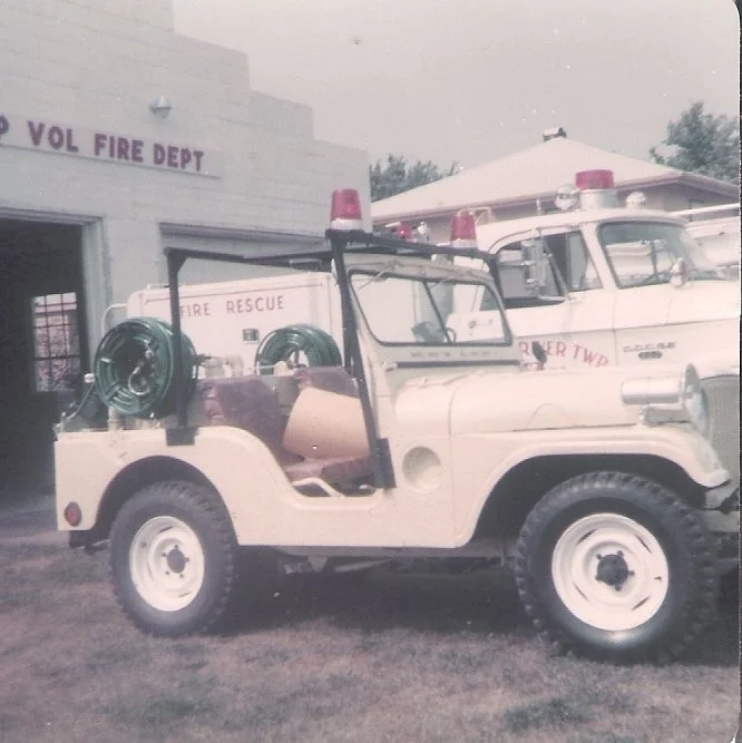 Vintage fire rescue vehicle parked outside a fire department building, with firefighting equipment and additional emergency vehicles visible.