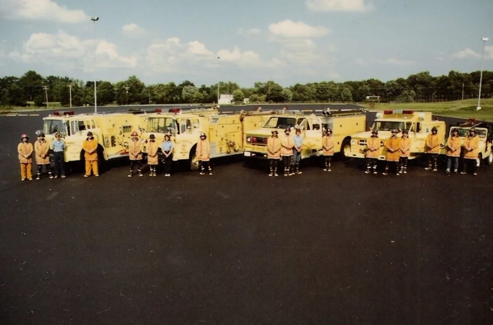 A group of children and firefighters standing in front of yellow fire trucks on a parking lot, with trees and a cloudy sky in the background.