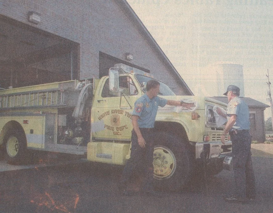 Two firefighters in uniform standing next to a yellow fire truck, discussing something outside a building.
