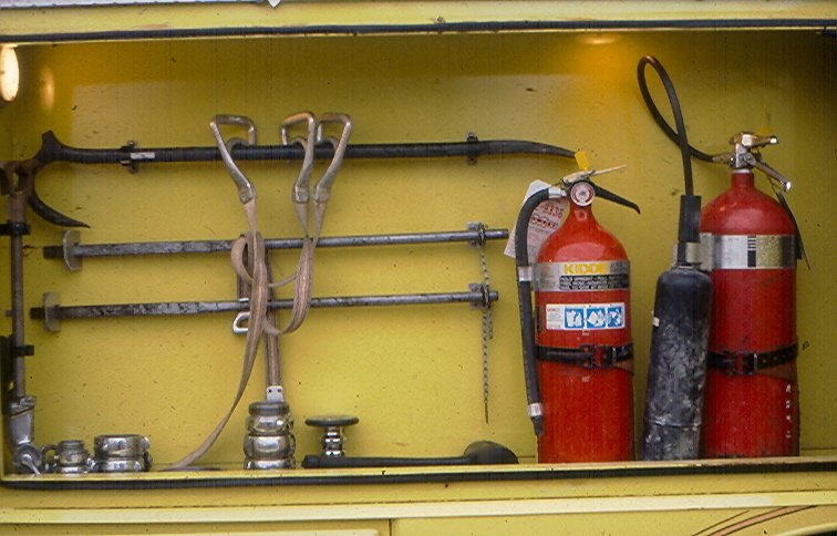 Fire extinguishers and various tools mounted on a yellow wall, including hooks, a chain, and hand tools.