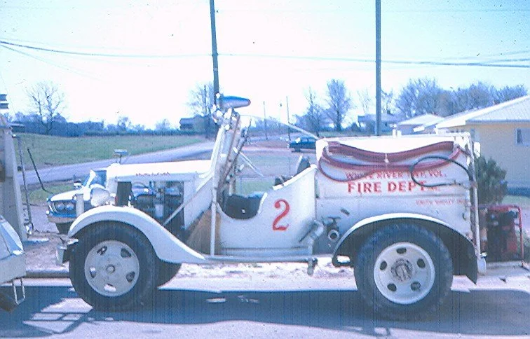 A white vintage fire truck with the number 2 on its side, parked outdoors near power lines and trees.