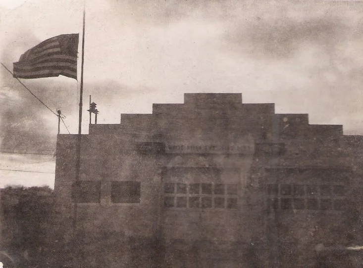 Black and white photo of a building with an American flag flying on a flagpole outside. The building has large windows and a stepped roofline.