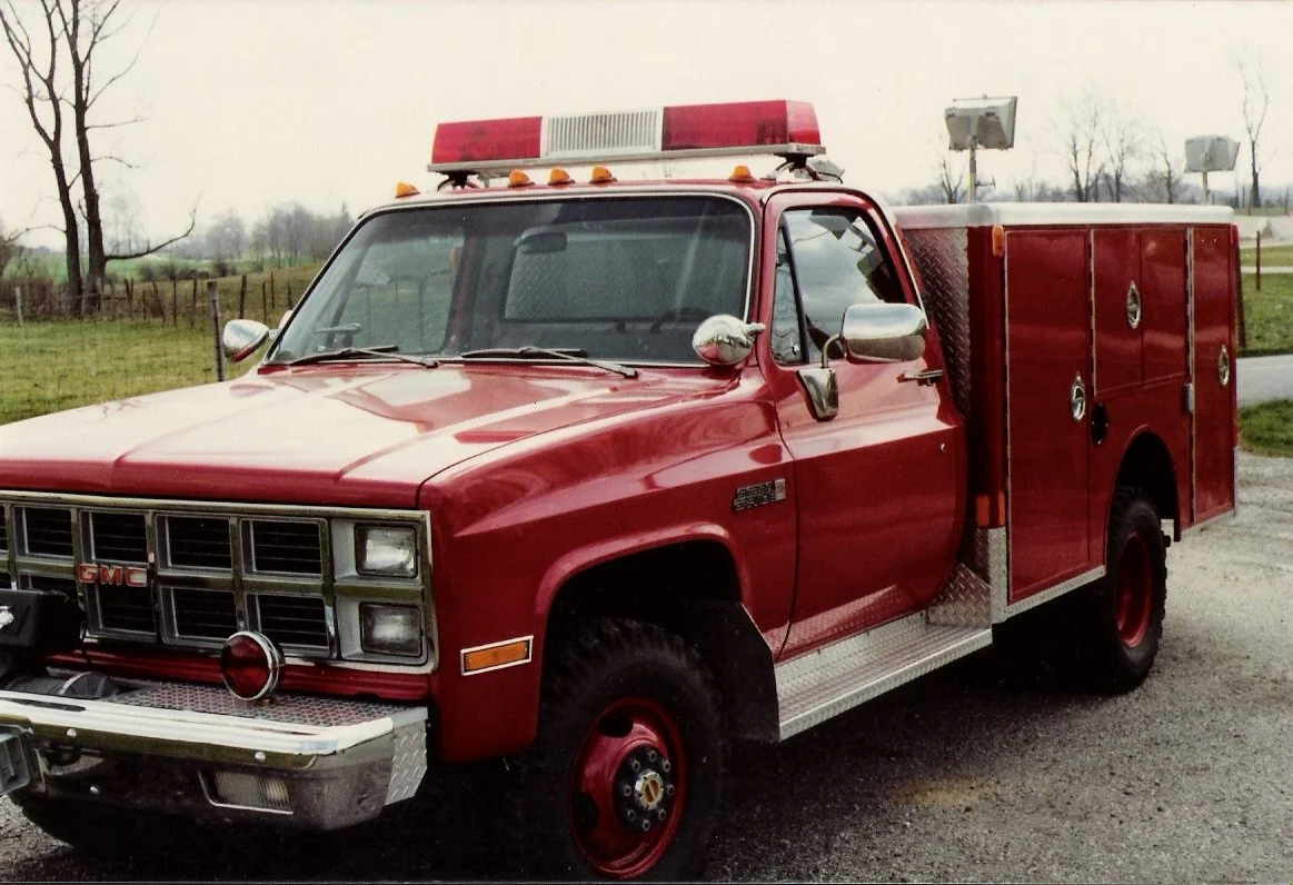 Red GMC emergency rescue truck with a light bar on top, parked on a gravel road near open fields with some trees in the background.