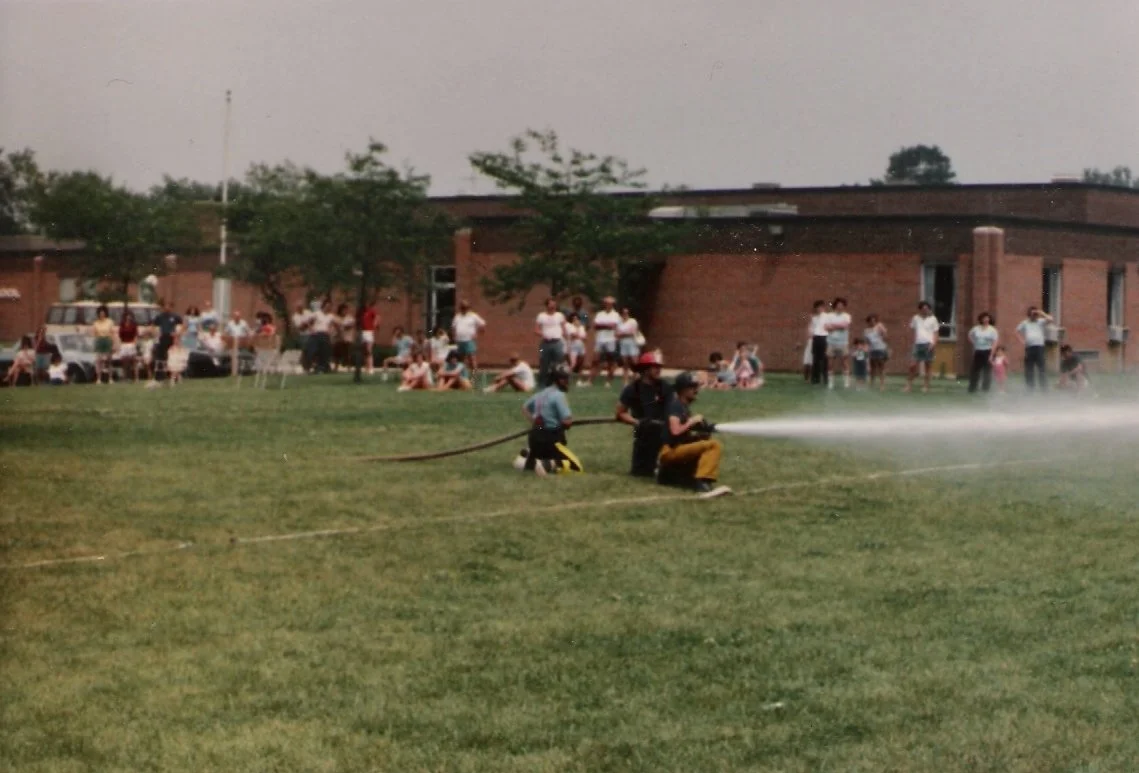 People gathered outside a brick building, watching firefighters practice with a fire hose on a grassy field during daytime.