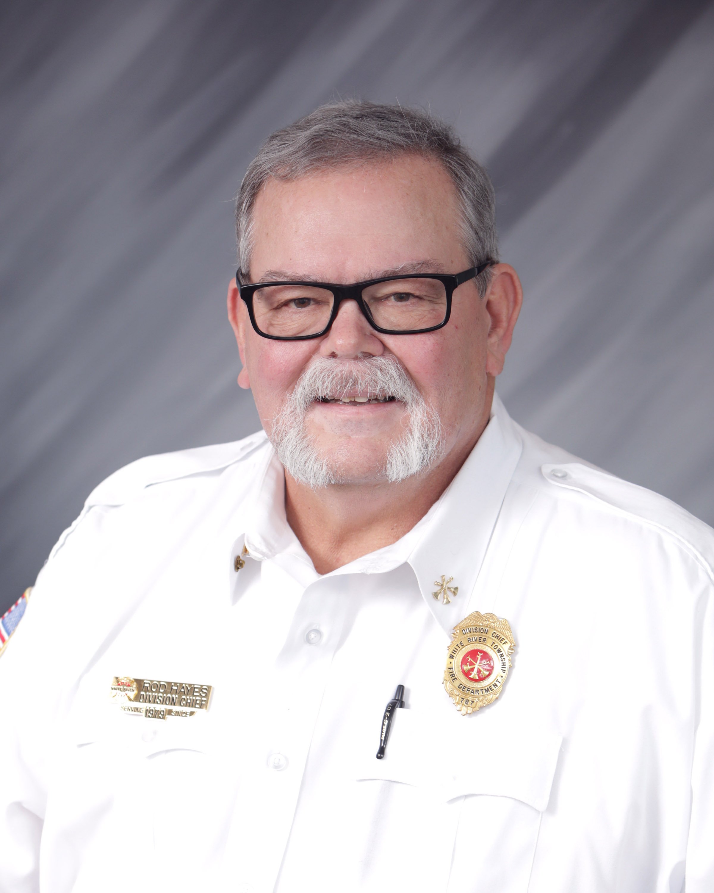 A man wearing a white uniform with fire department badges and patches, black glasses, gray hair, and a beard, smiling at the camera with a gray gradient background.