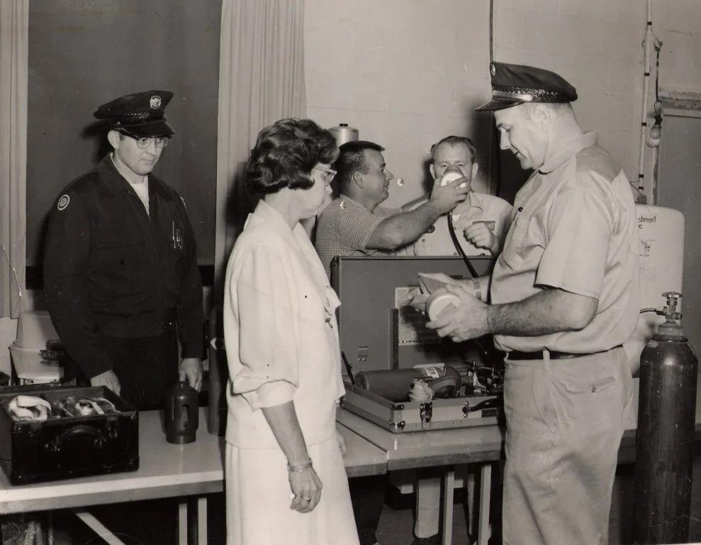 A group of emergency responders, including police officers and paramedics, gathered around a man in a uniform, examining medical equipment in a black-and-white photo.