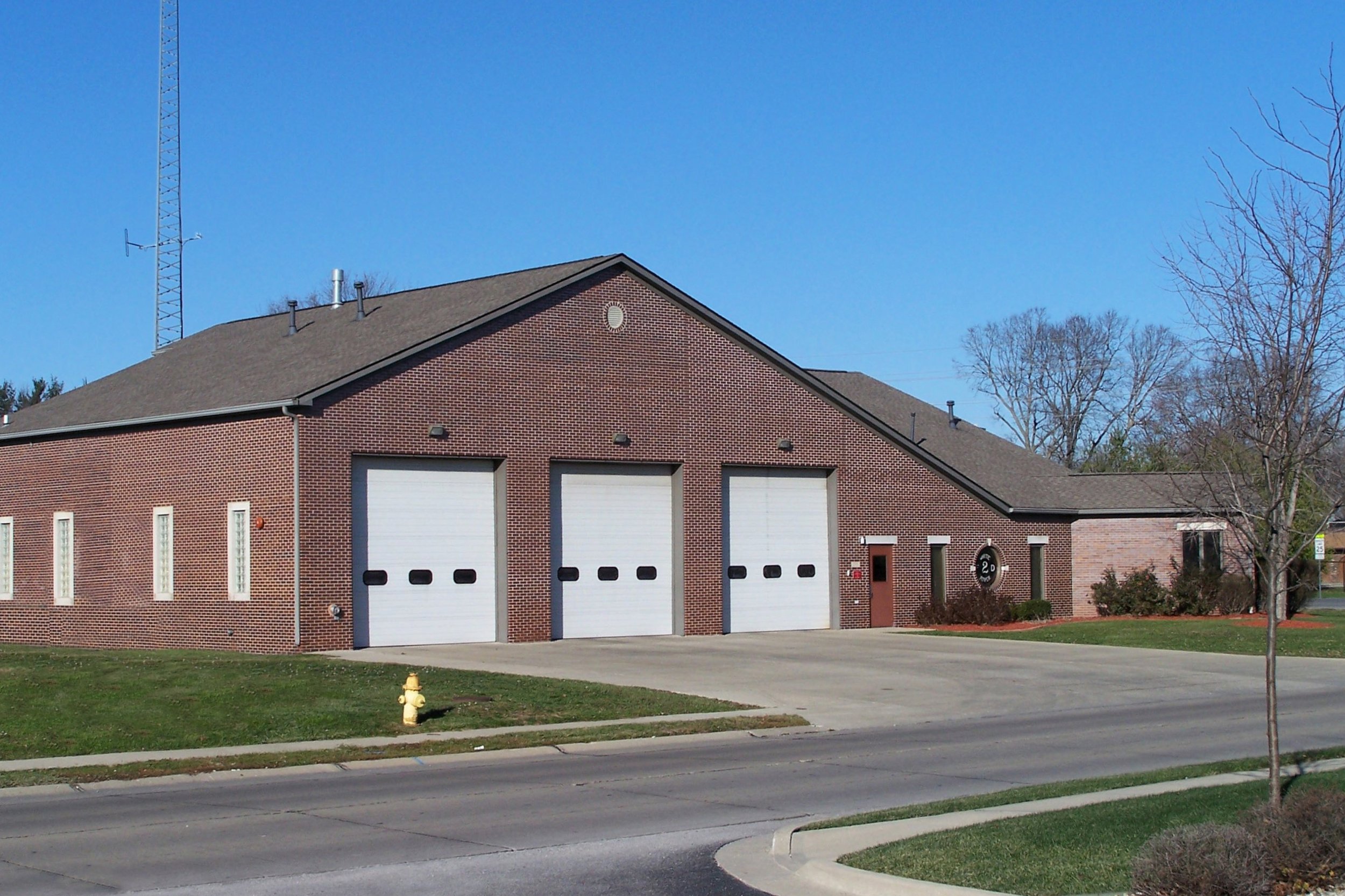 A brick building with three large white garage doors, a small door to the right with the number 20, and a fire hydrant on a grass patch in front. There is a leafless tree to the right and a tall antenna tower in the background under a clear blue sky.