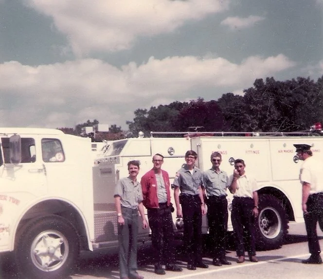 Five people standing in front of a large fire truck in an outdoor parking lot, with trees and a partly cloudy sky in the background.