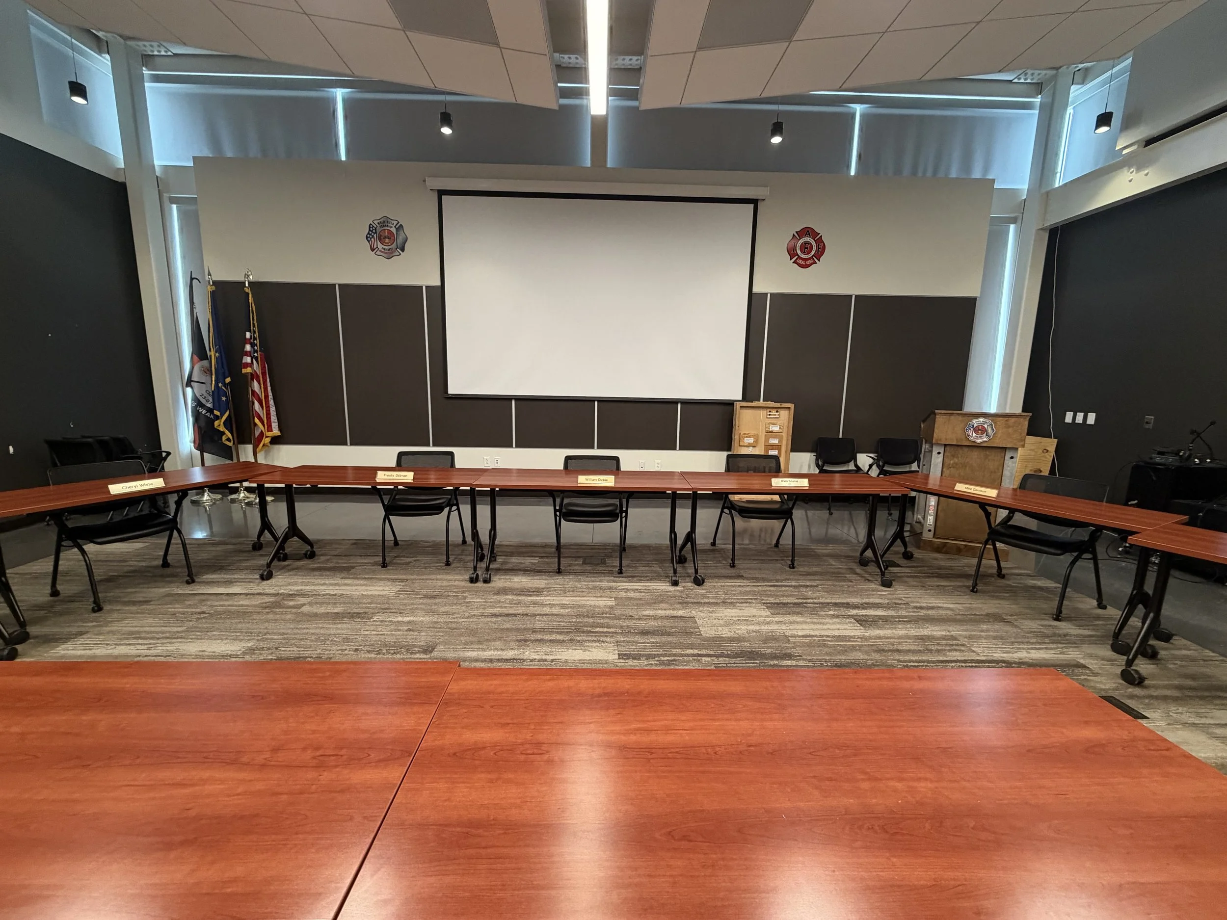 Empty conference room with U-shaped arrangements of wooden tables and black chairs, a large projection screen on the wall, flags on the left, and firefighter insignias on the wall.