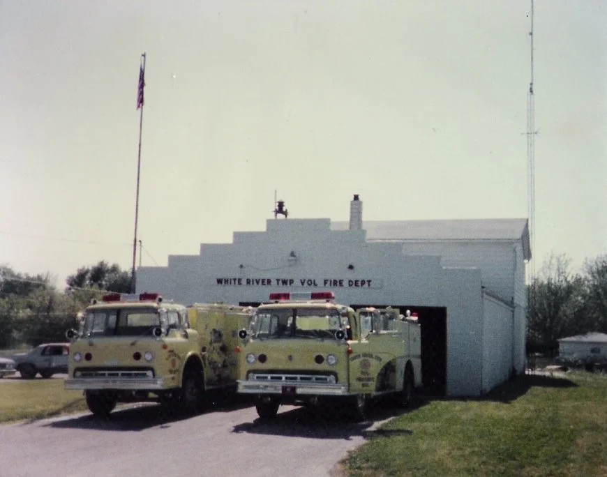 Fire station building with a sign reading "White River Twp Vol Fire Dept" and two vintage fire trucks parked in front.