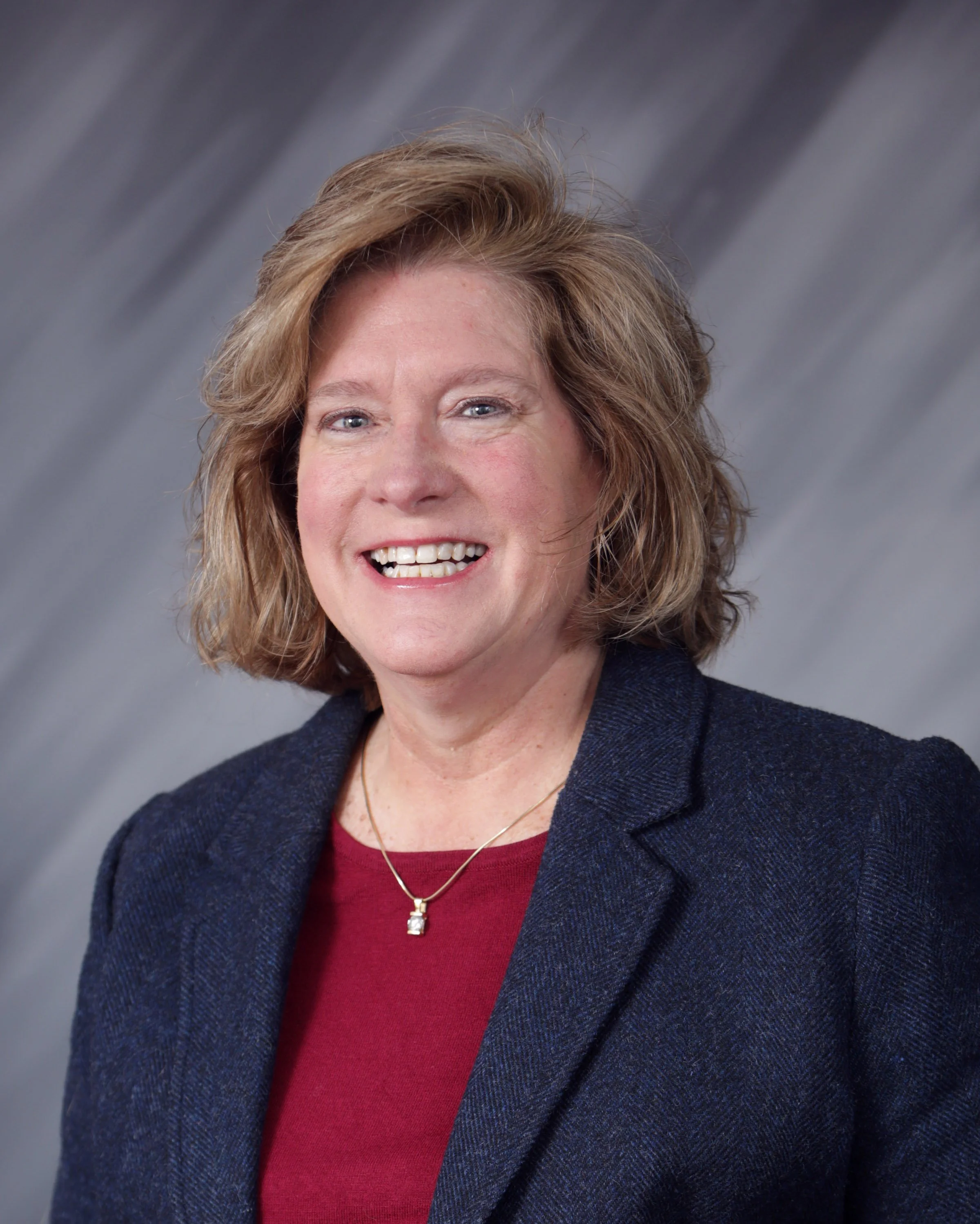 A smiling woman with shoulder-length light brown hair, wearing a navy blazer and red top, and a gold necklace, posing against a gray background.
