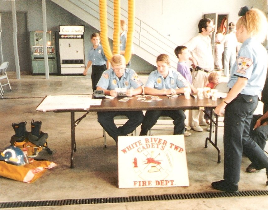 Young cadets at a fire safety event, sitting at a table with firefighting gear, with a sign reading  White River Twp. Cadets Fire Dept., inside a fire station or community center.