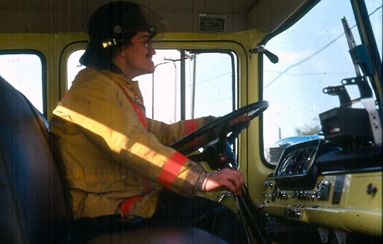 A person wearing a yellow and red jacket and a black helmet driving a large vehicle, possibly a truck, with the dashboard and steering wheel visible and large windows showing a clear sky outside.