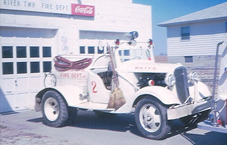 Vintage fire truck parked in front of a building with a Coca-Cola sign, labeled 'River Tov Fire Dept'.